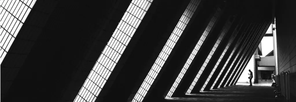 A person stands beneath a series of large, angled, sunlit beams in a modern architectural corridor.