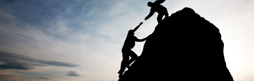 Two people climbing a steep rock formation, with one reaching down to help the other against a cloudy sky backdrop.