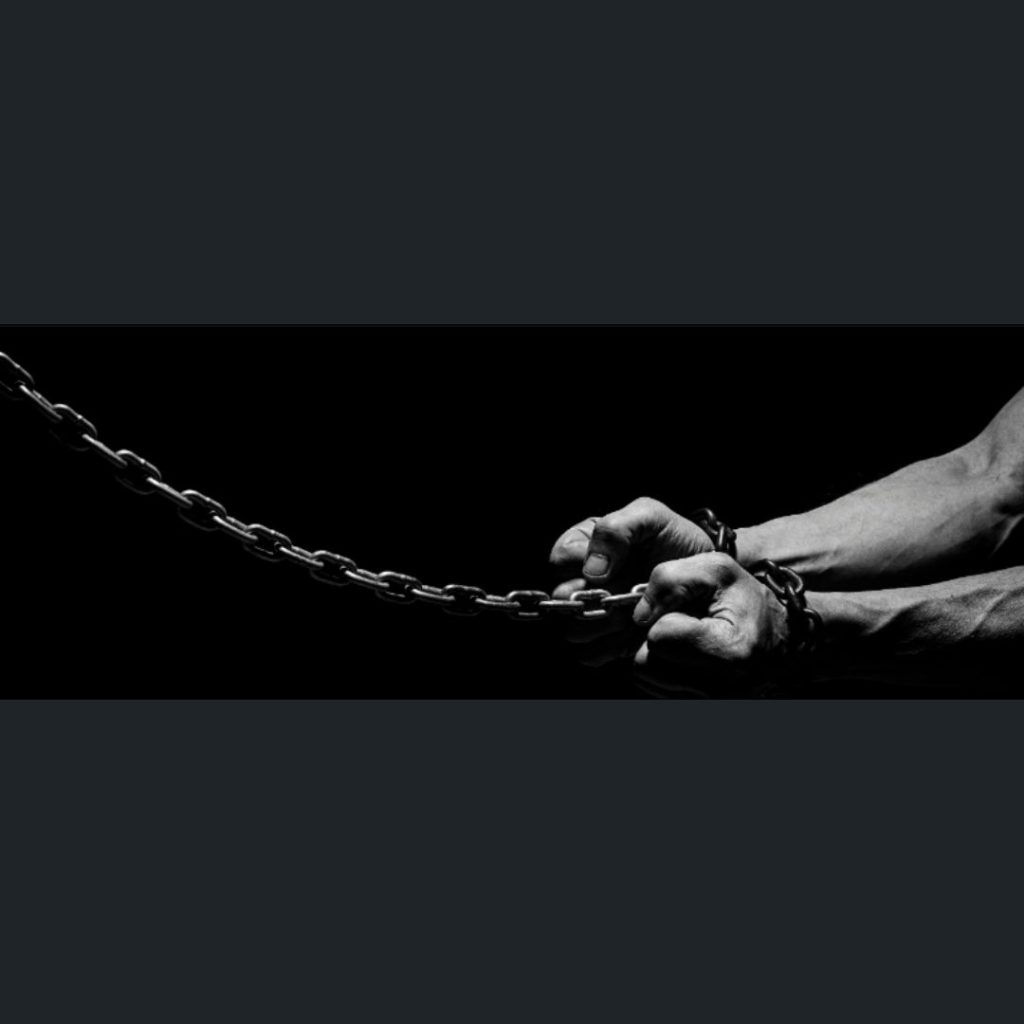 Two hands tightly gripping a heavy metal chain against a black background, with wrists appearing bound or restrained.