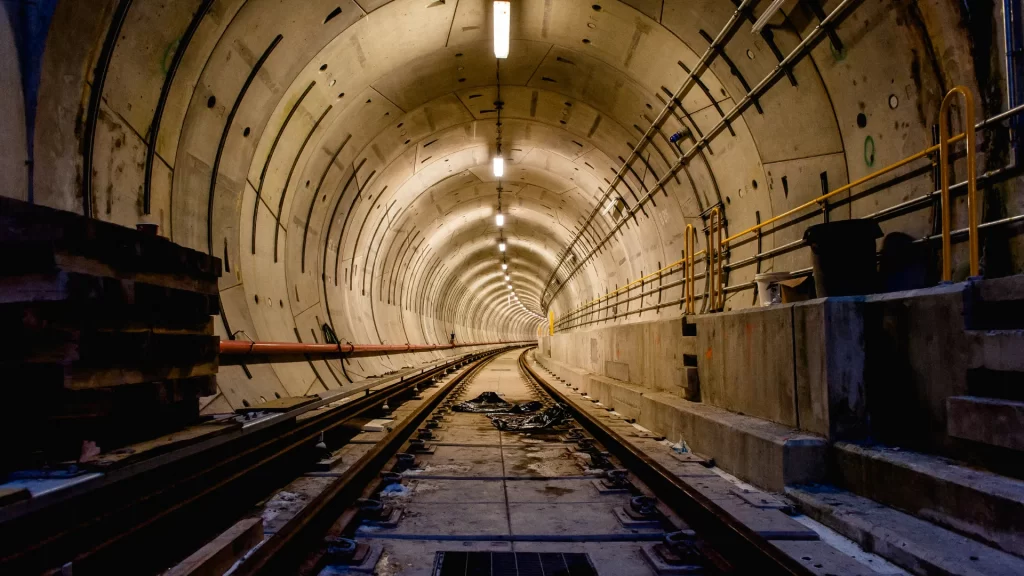 Underground tunnel with concrete walls, lit by overhead lights, featuring train tracks and construction materials along the sides.