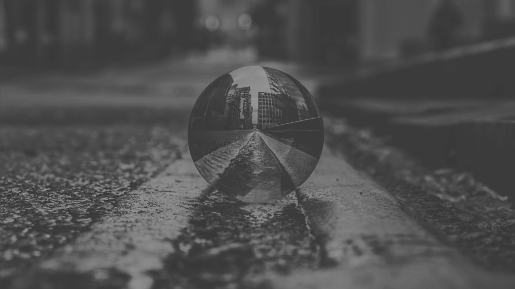 A glass sphere rests on a wet street, reflecting an upside-down image of surrounding buildings in a grayscale urban environment.