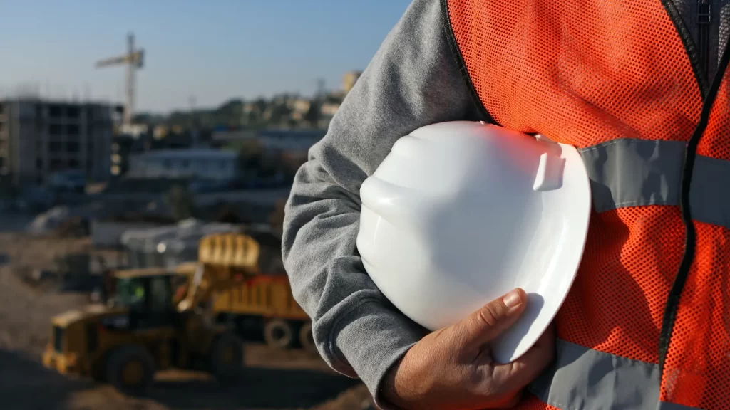 Person wearing an orange safety vest holds a white hard hat at a construction site, with construction vehicles and buildings visible in the background.