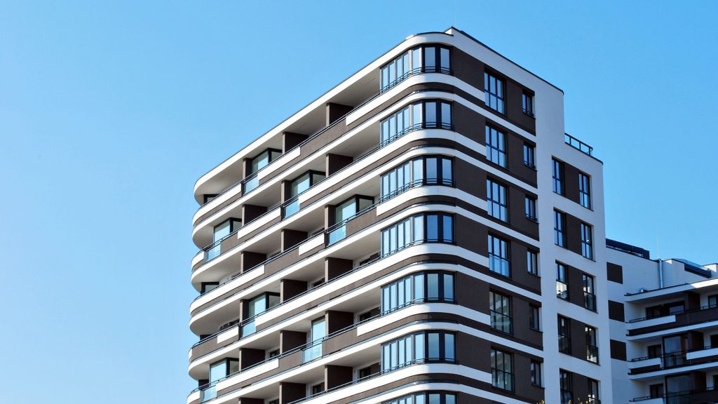 A modern multi-story apartment building with balconies, large windows, and a geometric facade, set against a clear blue sky.