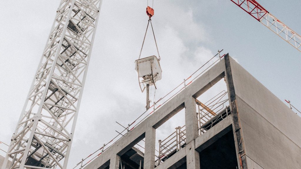 A crane lifts construction materials beside a partially built concrete building under a cloudy sky.
