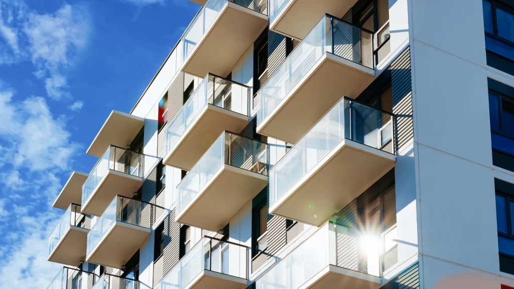 Modern apartment building with multiple balconies and glass railings, viewed from below against a bright blue sky with scattered clouds. Sunlight reflects off one of the windows.