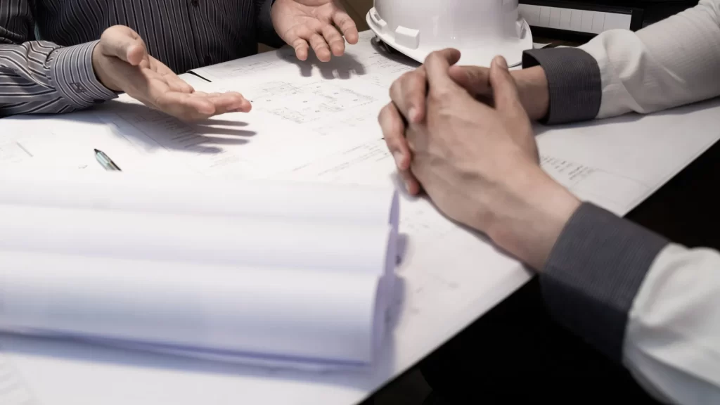Two people sit at a table with architectural blueprints and a white hard hat, discussing plans with hands visible.