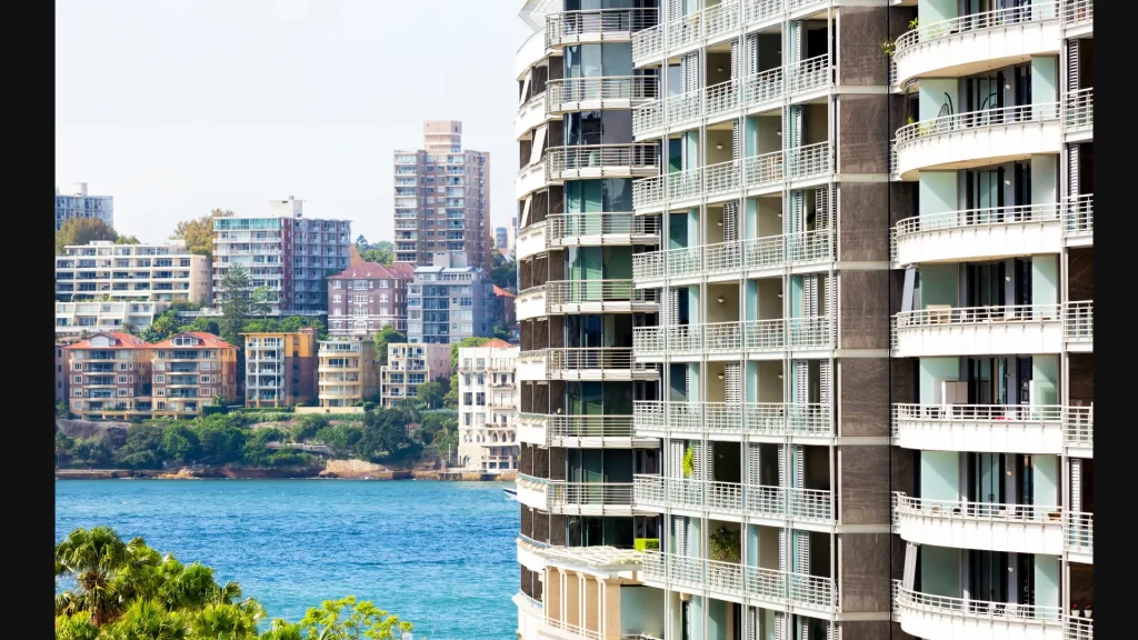 Modern waterfront apartment buildings with balconies overlook a blue body of water, with other residential buildings visible in the background.