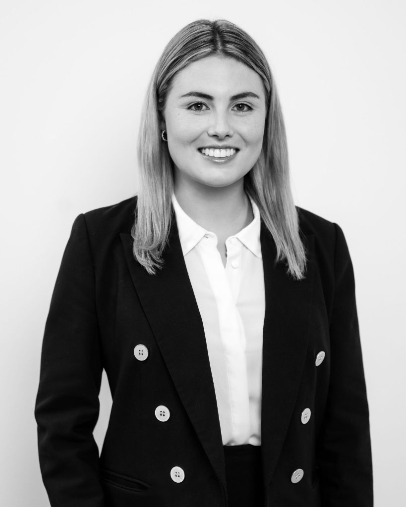 A woman with straight, shoulder-length hair wears a dark blazer over a light-colored blouse and smiles at the camera against a plain background. The image is in black and white.
