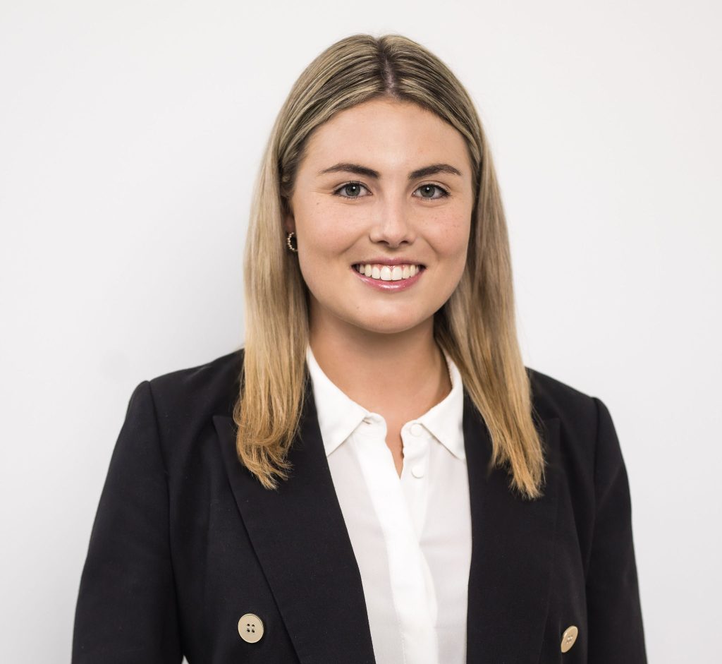A woman with straight blonde hair, wearing a black blazer over a white blouse, smiles at the camera against a plain white background.