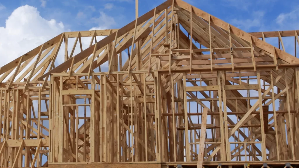 Wooden frame of a house under construction, with exposed beams and trusses set against a blue sky with some clouds.