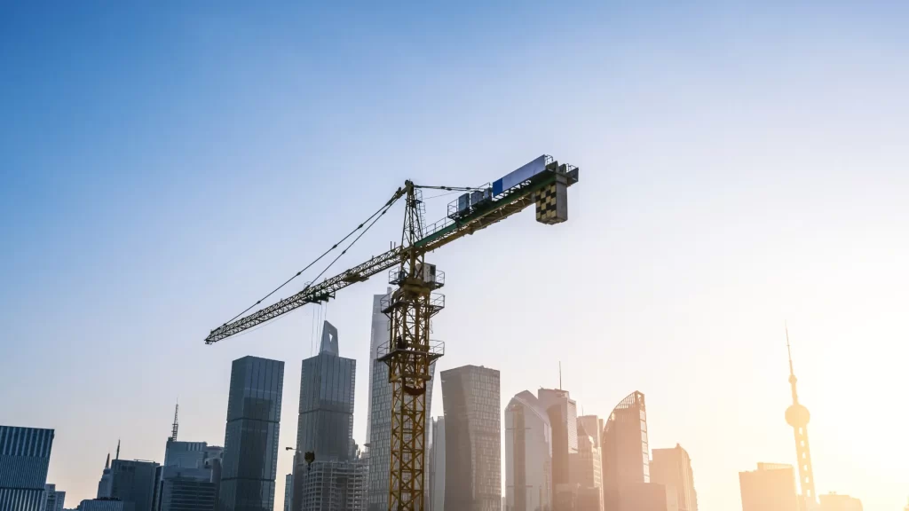 A construction crane stands in front of modern city skyscrapers under a clear blue sky at sunrise or sunset.