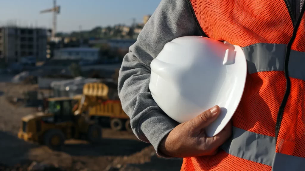A person wearing an orange safety vest holds a white hard hat at a construction site with heavy machinery and buildings in the background.