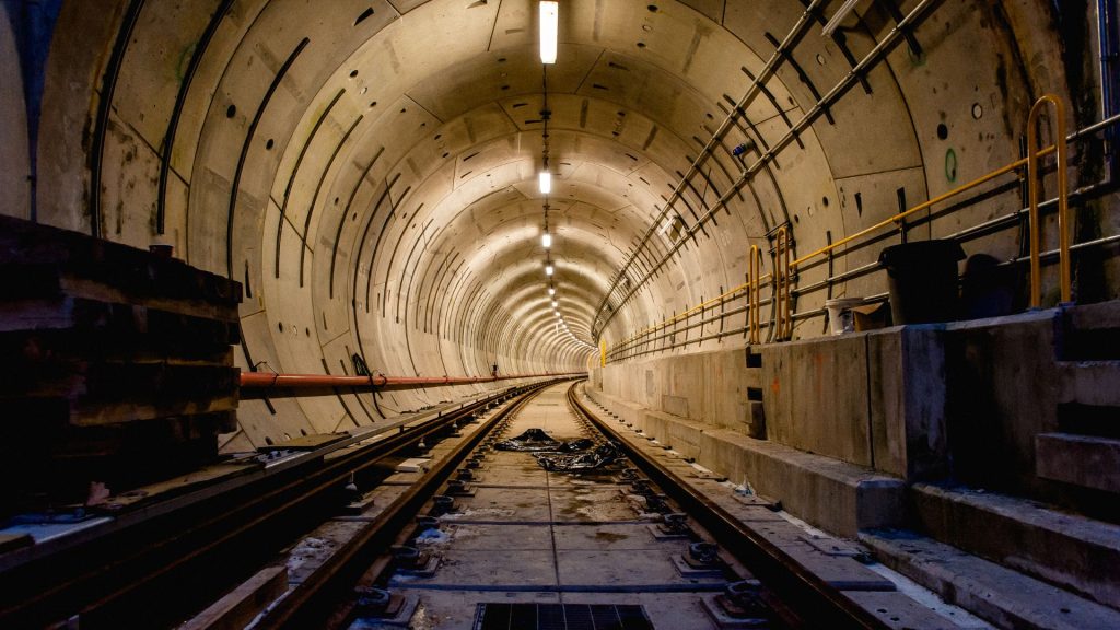 A well-lit, unfinished subway tunnel with metal tracks, concrete walls, and construction materials along the sides.