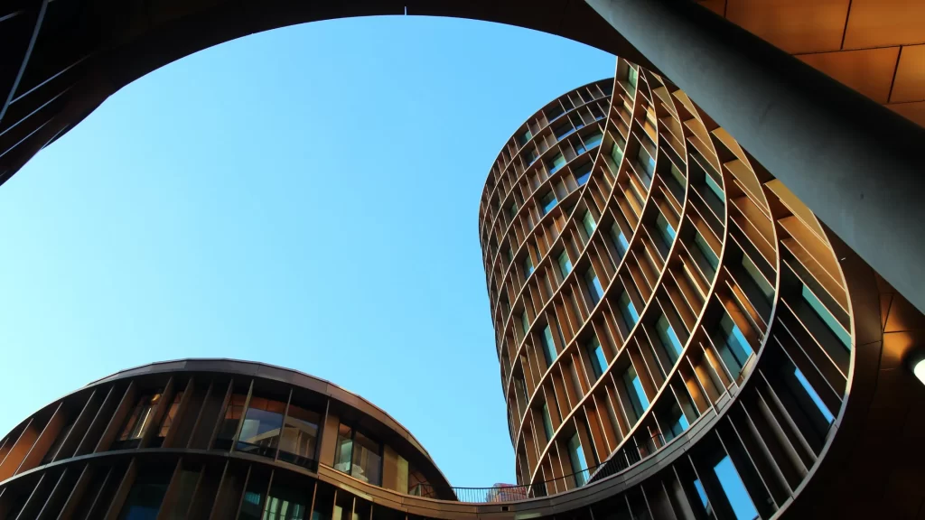 View looking up at modern, curved buildings with a pattern of brown and glass windows, framed by a circular architectural structure against a clear blue sky.