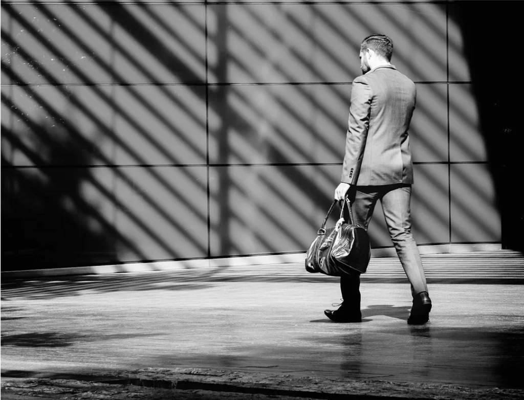 A man in a suit walks across a sunlit pavement, carrying a large bag, with diagonal shadows cast on a tiled wall behind him.