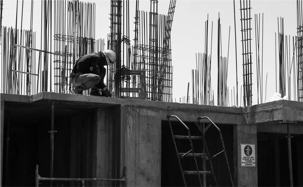 A construction worker wearing a helmet crouches on a concrete structure surrounded by vertical rebar at a building site.