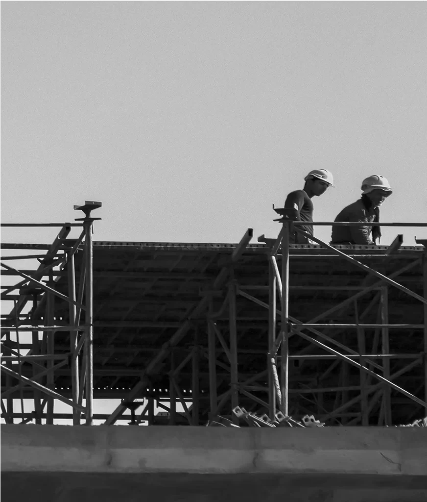 Two construction workers wearing hard hats stand on scaffolding at a building site, with a clear sky in the background.