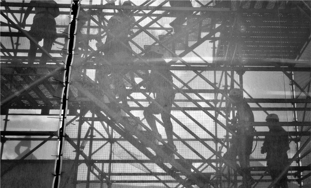 Silhouettes of construction workers are seen through mesh netting as they climb and stand on metal scaffolding.