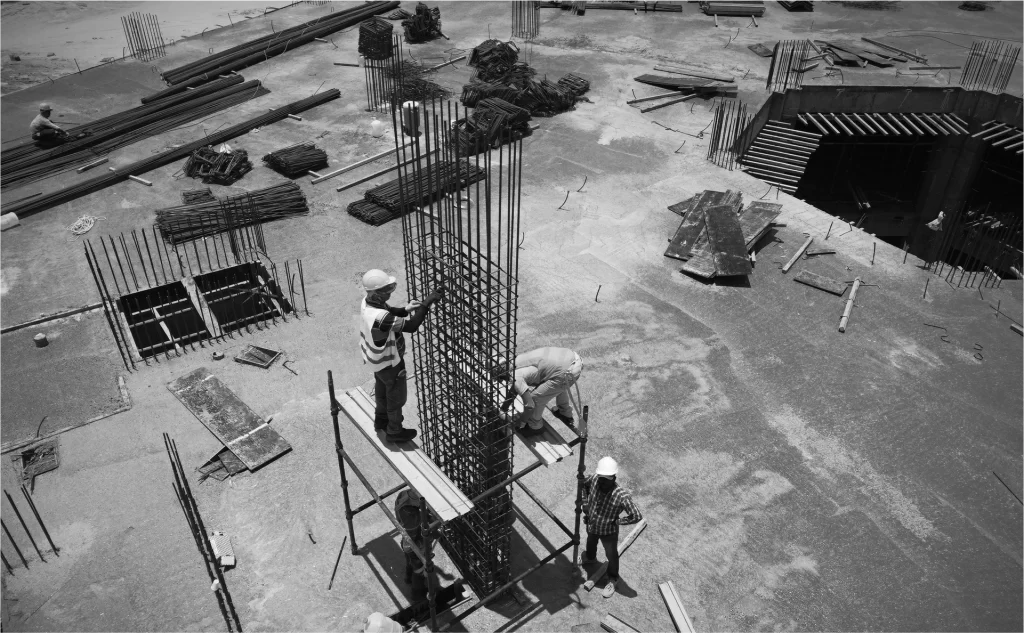 Construction workers assembling steel reinforcement bars at a building site; some stand on scaffolding, while others work on the ground amid scattered materials.