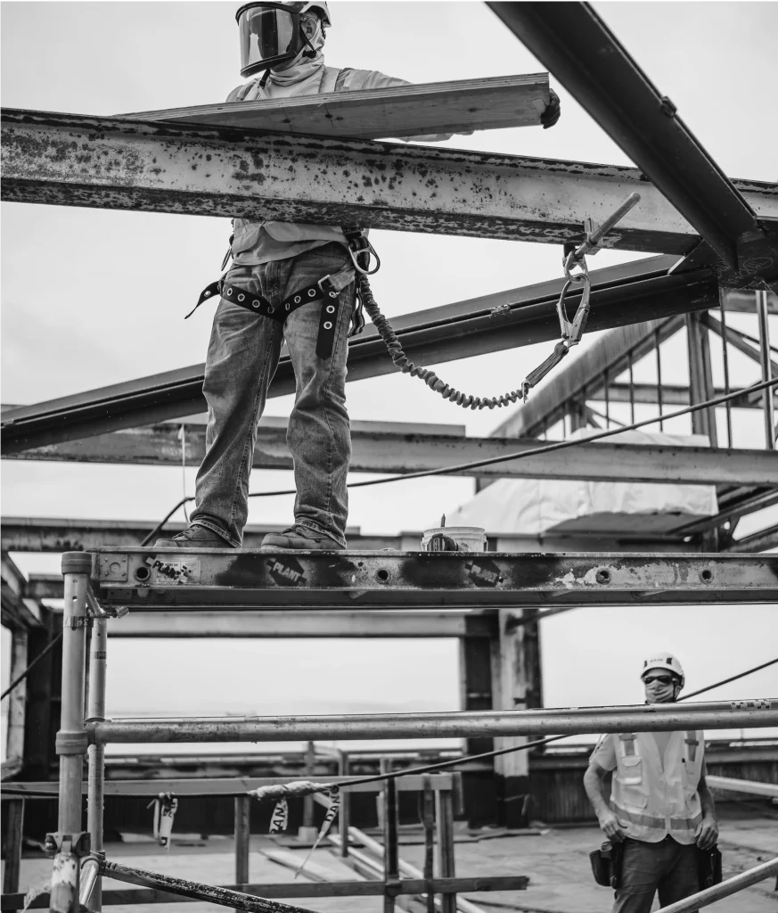 A construction worker in safety gear stands on a steel beam, secured by a harness, while another worker looks on from the ground at a building site.