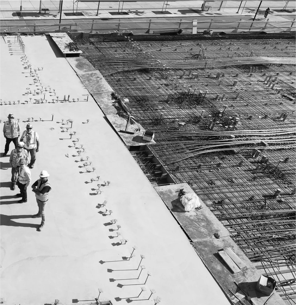 A group of construction workers in safety gear stand on a concrete foundation next to exposed steel rebar at a building site.