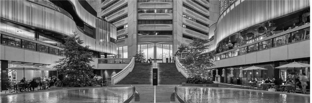 Black and white photo of a modern indoor atrium with a central staircase, water features, trees, tables with umbrellas, and multi-story balconies.