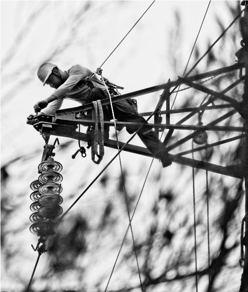 A utility worker wearing safety gear repairs electrical equipment while seated on a high power line structure.