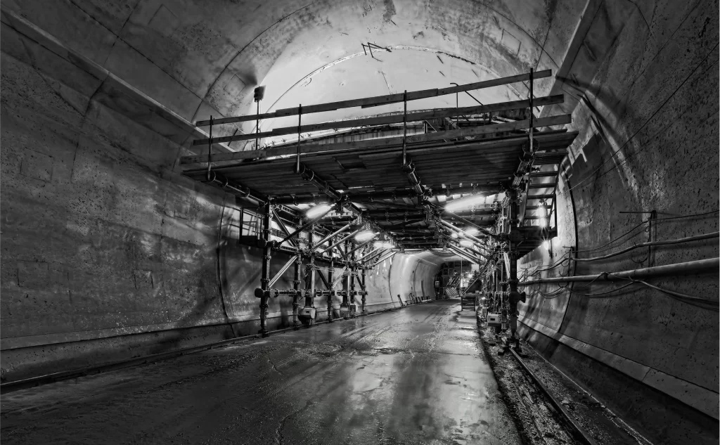 A large concrete tunnel with scaffolding and support structures overhead; cables and pipes run along the tunnel walls and the floor appears wet.