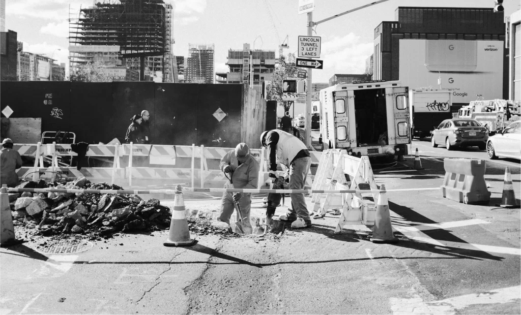 Two construction workers use jackhammers on a city street, surrounded by traffic cones and barriers, with buildings and an ambulance in the background.