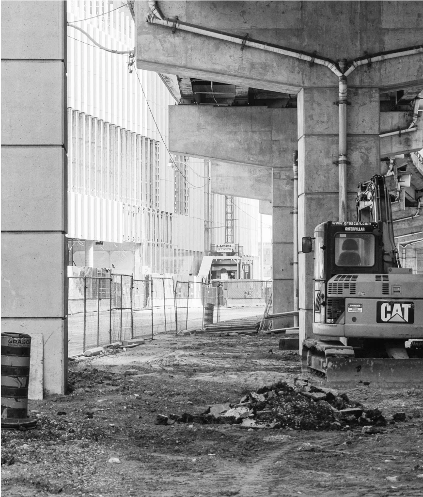A construction site under a concrete overpass with a CAT excavator, fencing, and debris visible on the ground.