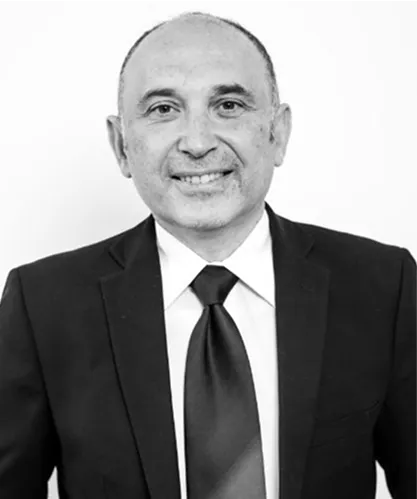 A man wearing a dark suit, white dress shirt, and dark tie is smiling at the camera against a plain, light background.